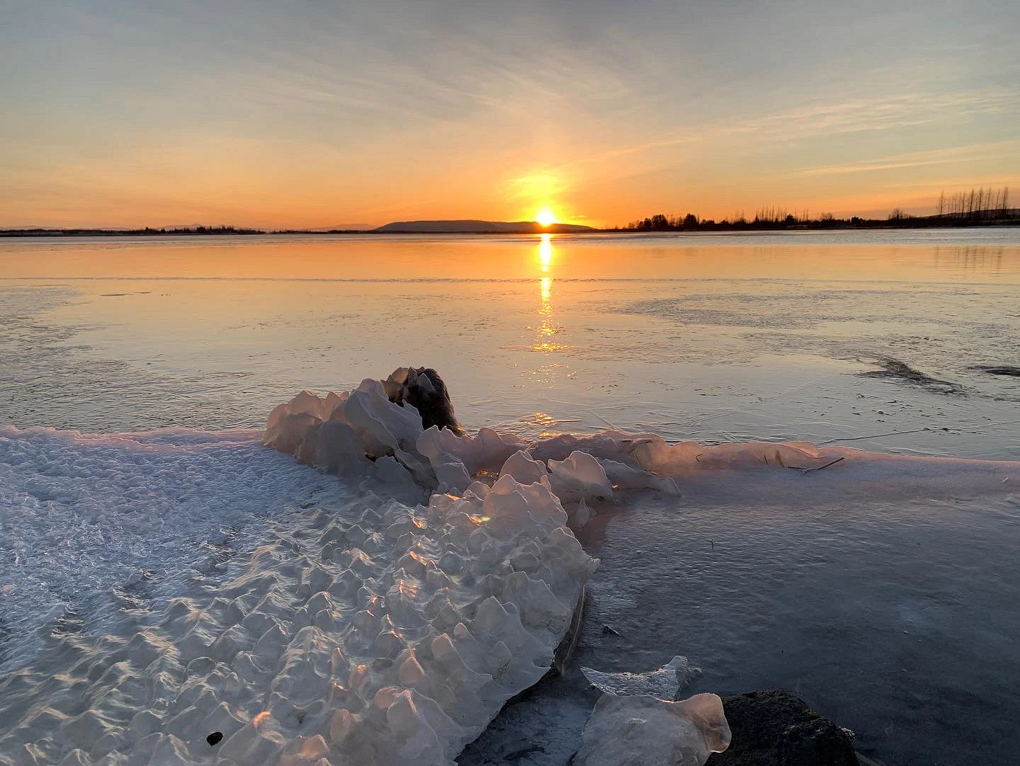 Frozen shoreline with delicate ice formations and a golden sunset reflecting on the calm water.